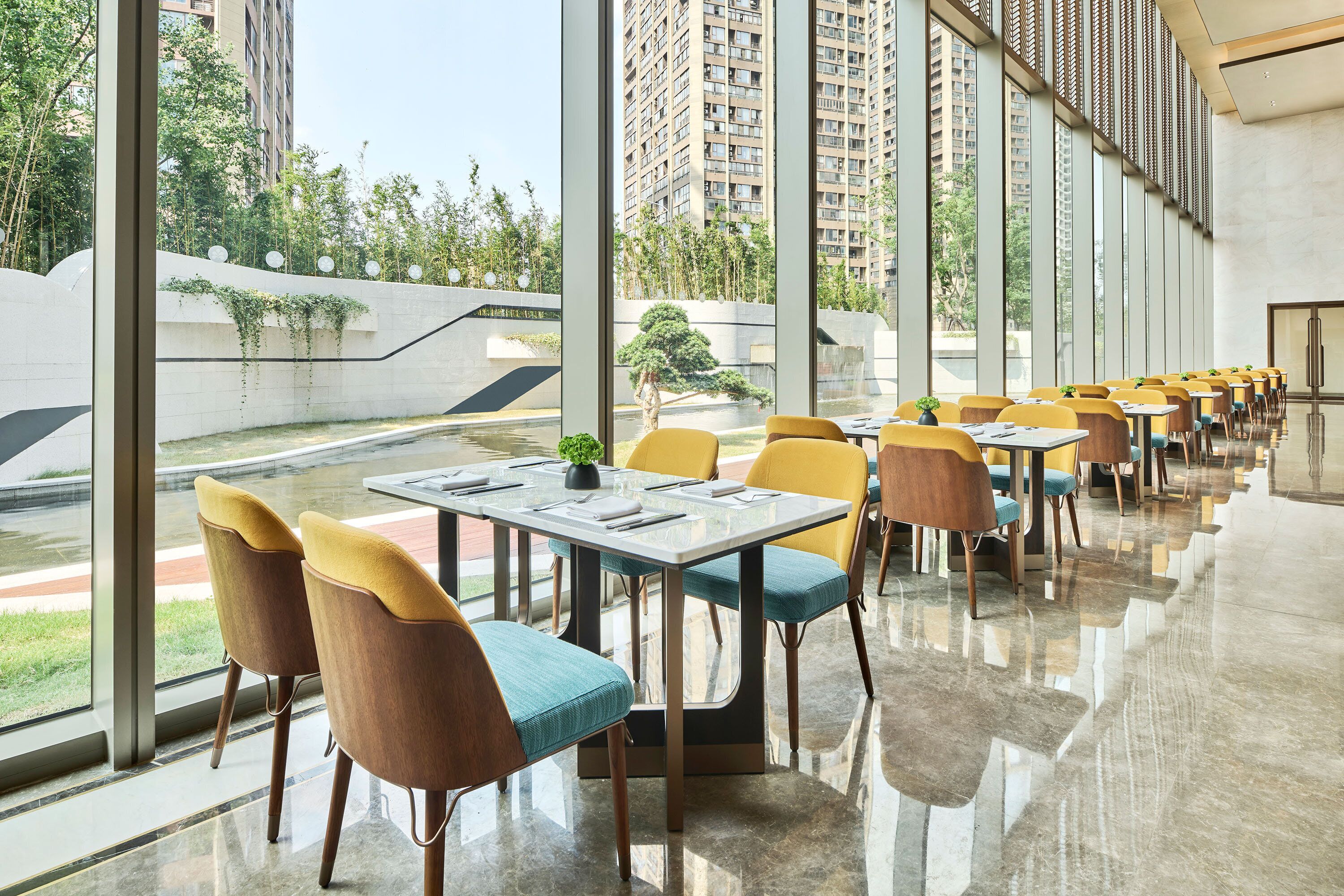 A modern dining area with tables and chairs arranged in rows, large windows offering views of a landscaped area and buildings.