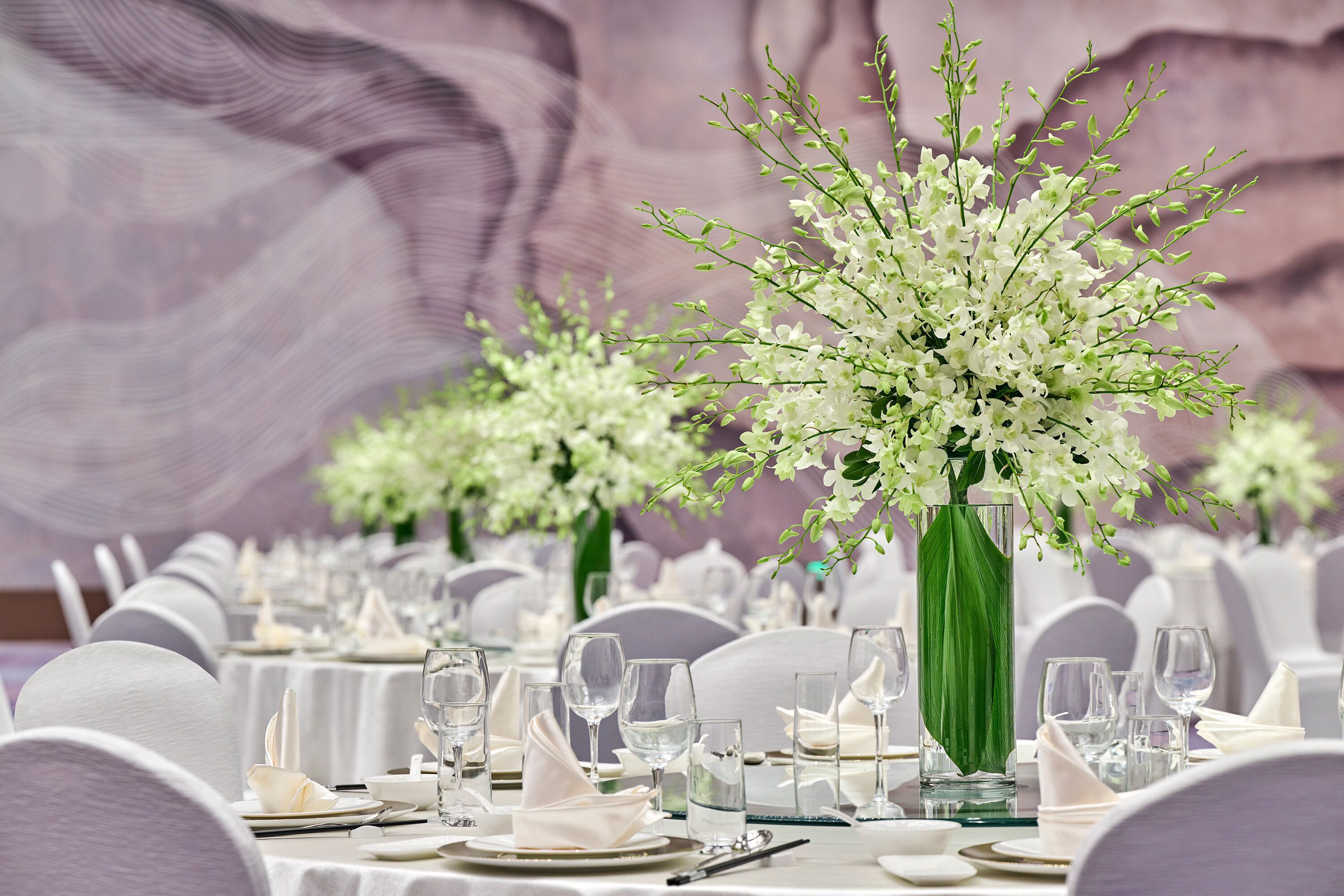 Una mesa de banquete decorada con manteles blancos, cristalería y un gran centro de mesa floral blanco.