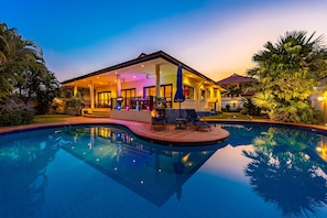 Un espace aménagé au bord de la piscine, comprenant un coin salon couvert, une piscine et une végétation luxuriante.