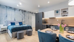Ein modernes Schlafzimmer mit Bett, Nachttischen und einem kleinen Sitzbereich mit Sessel und Hocker. Im Zimmer befindet sich ein großes Fenster und eine Wand mit Baumdekor.