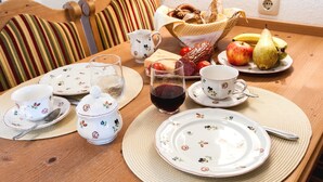 Uma mesa de jantar de madeira posta com louças de padrão floral, um cesto de pão, frutas e uma jarra de bebida vermelha.