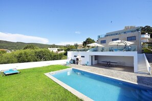A modern house with a swimming pool, outdoor seating, and a glass railing overlooking a green landscape.