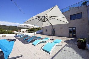 A rooftop terrace with sun loungers and large umbrellas, a building with a modern design, and a view of the surrounding landscape.