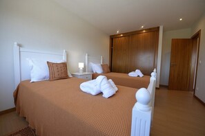 A hotel room with two beds, each with white headboards and brown bedding. There are white towels on the beds, and a wooden wardrobe is visible in the background.