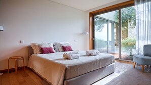 Een slaapkamer met een bed, een klein krukje, een stoel en een grote glazen schuifdeur die leidt naar een buitenruimte.