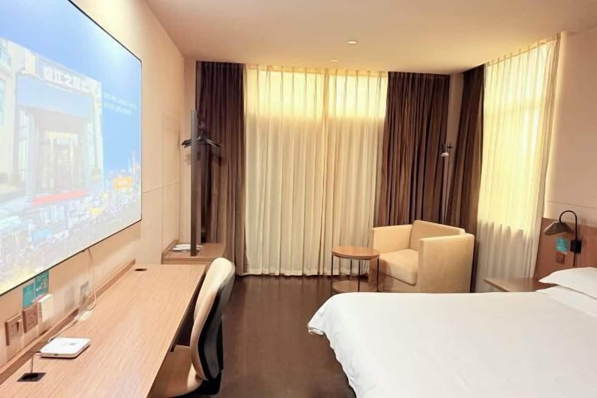 A hotel room with a bed, a desk with a chair, a small table, and a projector screen displaying a cityscape.