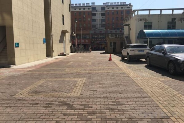 Una zona pavimentada con la entrada de un edificio, un toldo azul y coches aparcados.