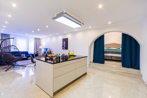 A modern hotel room with a kitchen area, a seating area with a sofa and chair, and a bedroom with a bed and curtains.