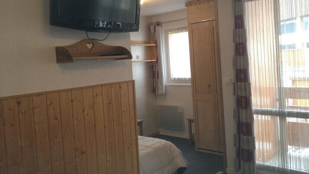 A bedroom with a bed, a television mounted on the wall, a wooden headboard, a window with curtains, and a radiator.