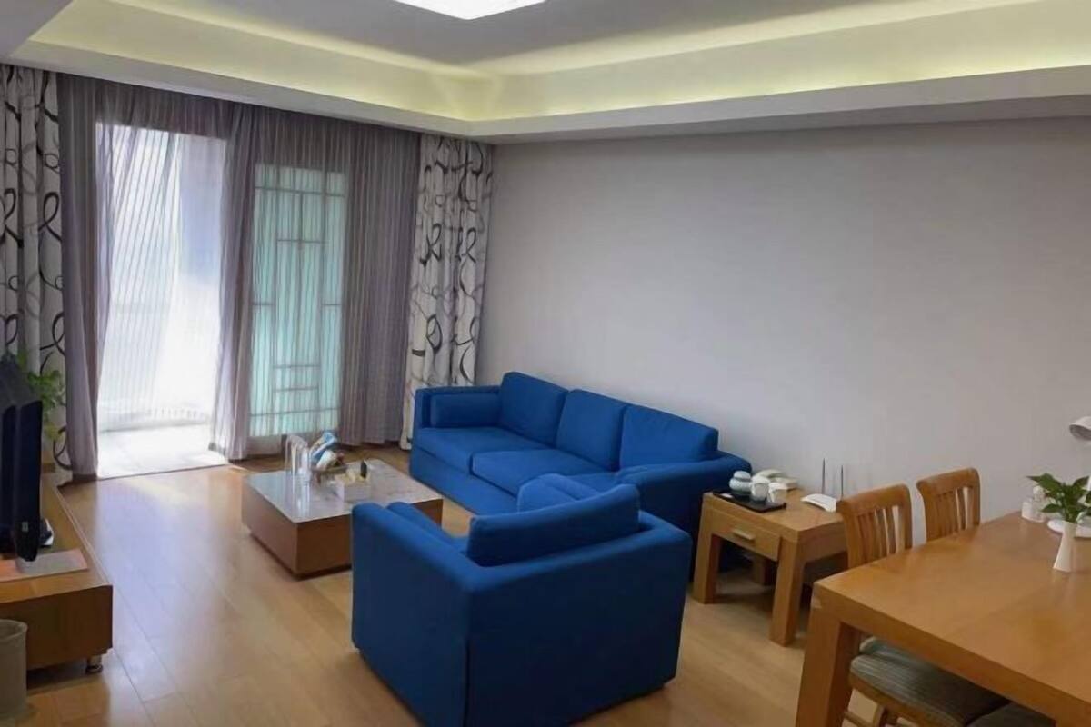 Ein modernes Wohnzimmer mit einem blauen Sofa, einem Couchtisch, einem Esstisch mit Stühlen und einem großen Fenster mit Vorhängen.