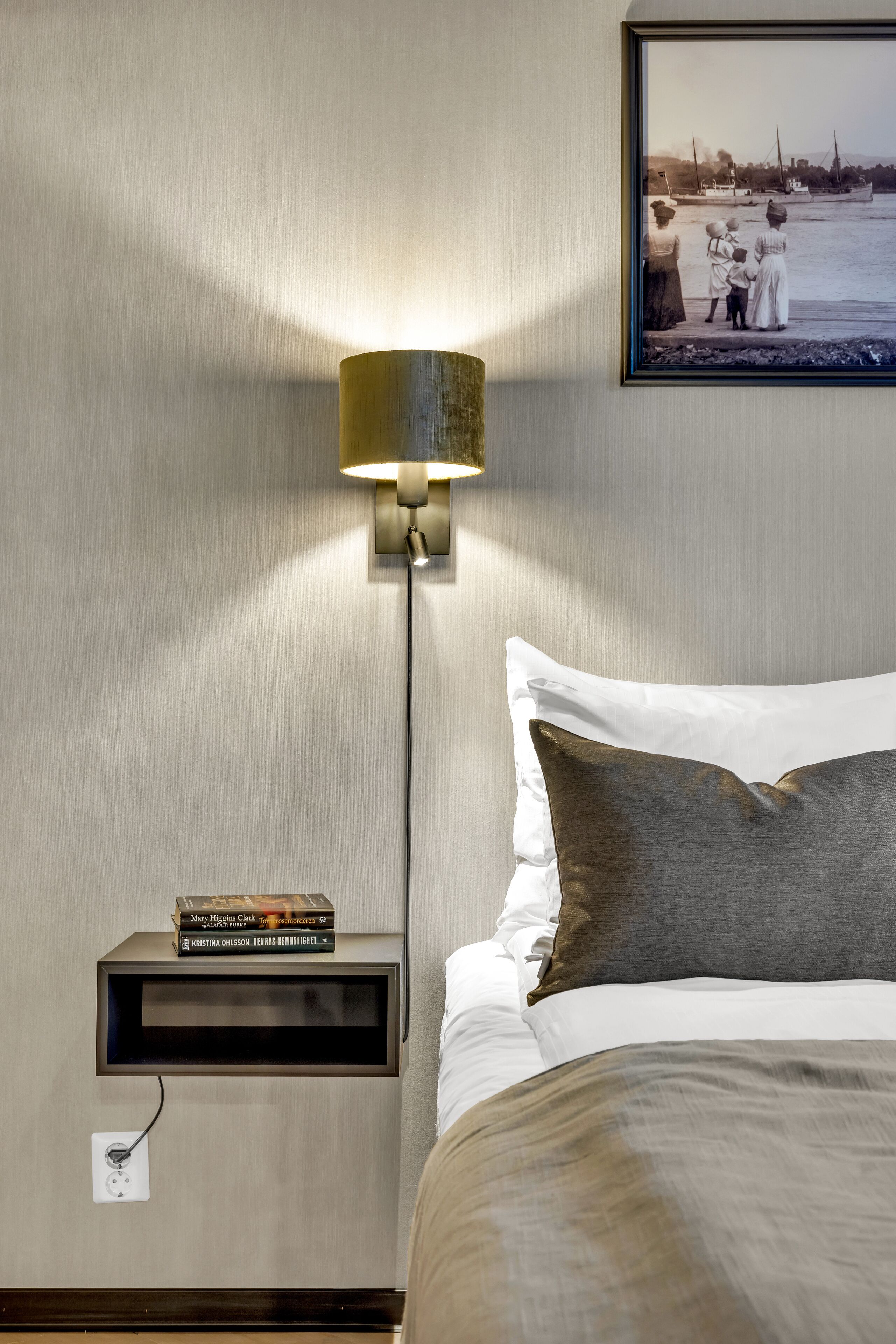 A neatly made bed with a white and brown pillow, a bedside table with a lamp, and a framed photo on the wall.
