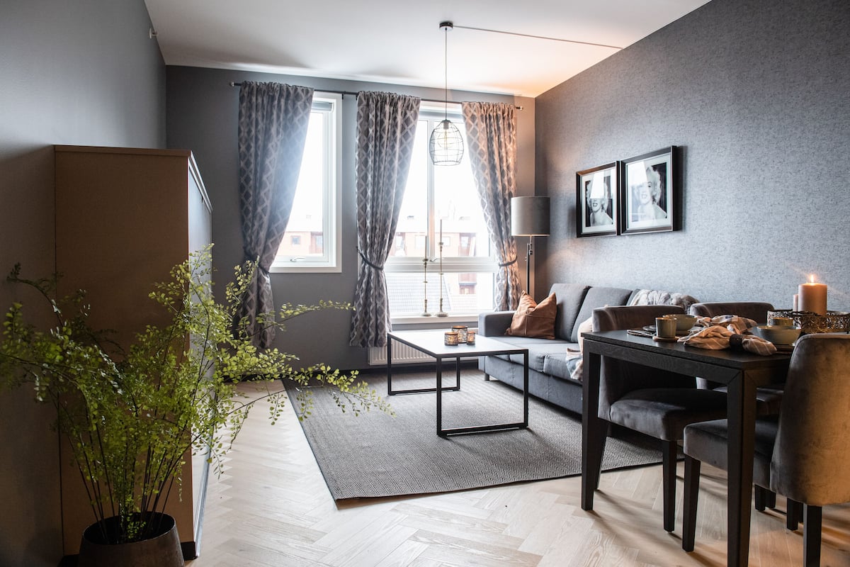 Un salon moderne avec un canapé, une table basse et des fauteuils. On y trouve une grande fenêtre avec des rideaux, une plante en pot et une lampe.