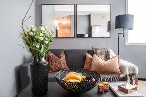 A modern living room with a grey sofa, decorative pillows, a black woven bowl with fruits, a vase with white flowers, and a floor lamp. - Origo Leilighetshotell  (Drammen)