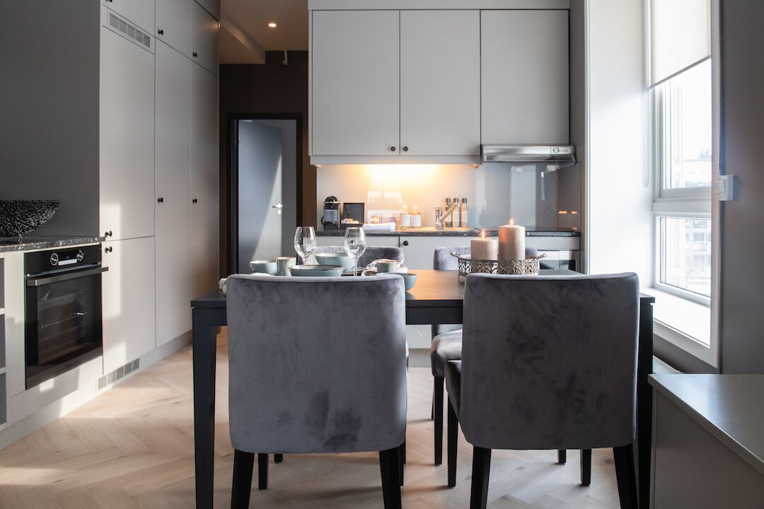 A modern kitchen with white cabinets, a built-in oven, and a dining area with two grey upholstered chairs.