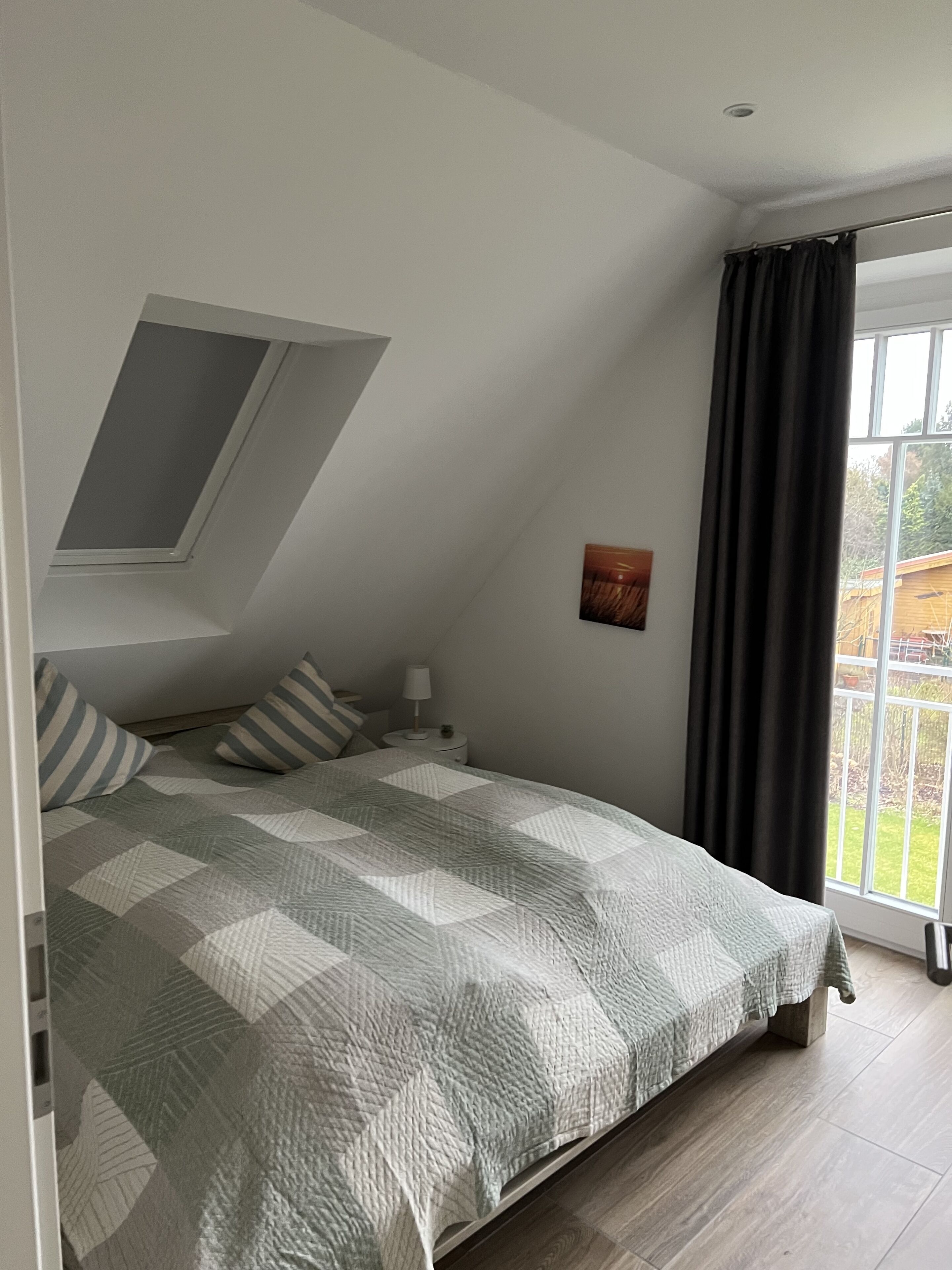 A bedroom with a sloped ceiling, a bed with striped pillows, a bedside table with a lamp, and a window with a view of the outdoors.