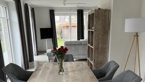 A dining area with a wooden table, grey upholstered chairs, a vase with red flowers, and a television in the background. - Spacious vacation home near the beach in a quiet location in Rosenhagen (Dassow)