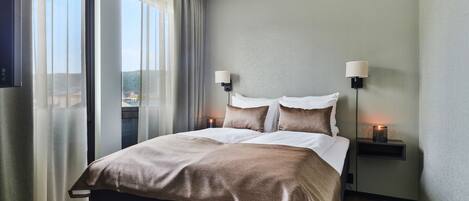 A modern hotel room with a large bed, two bedside lamps, and a view through the window.