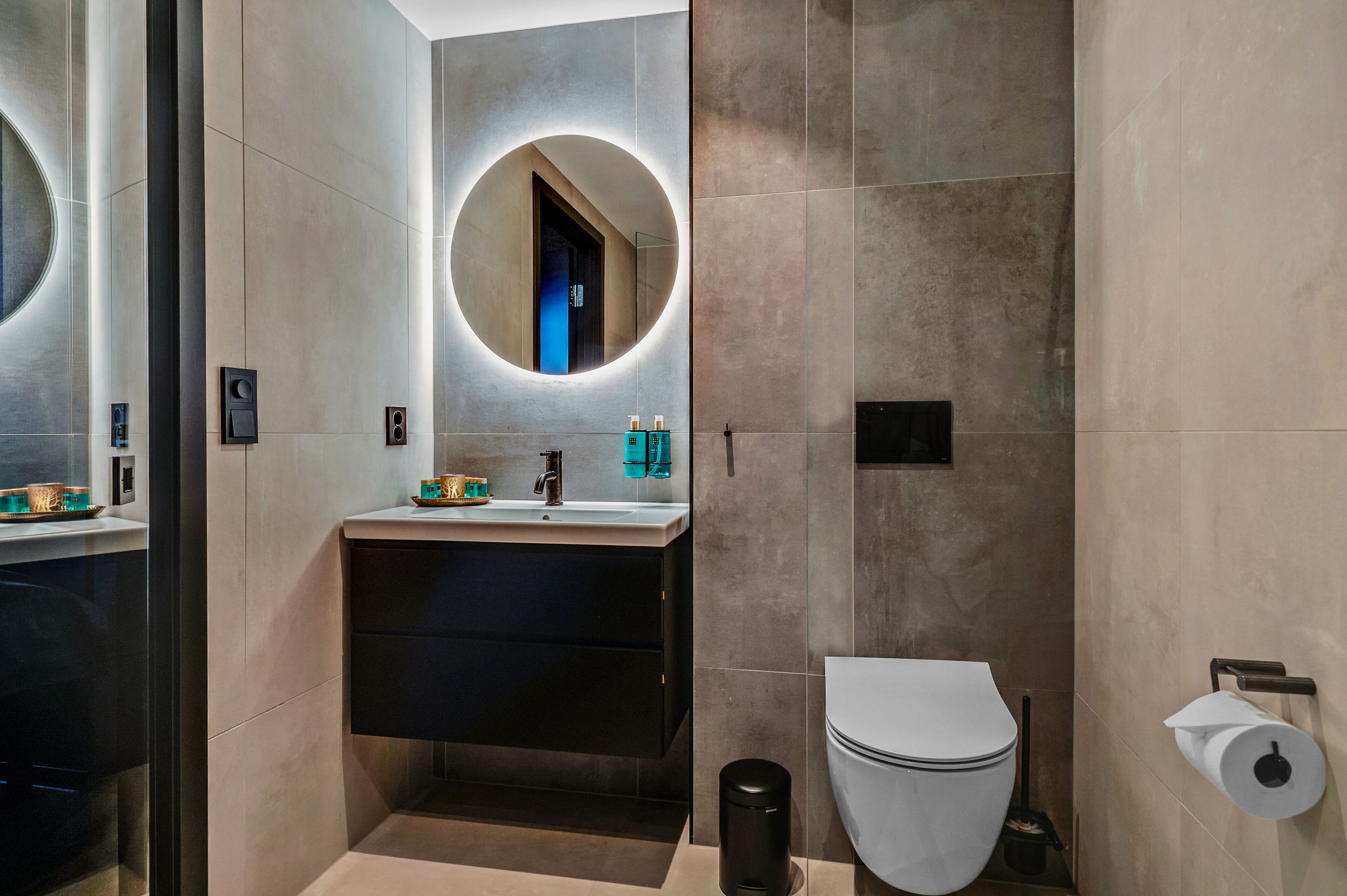 A modern bathroom with a round mirror, a sink, a toilet, and a trash bin.
