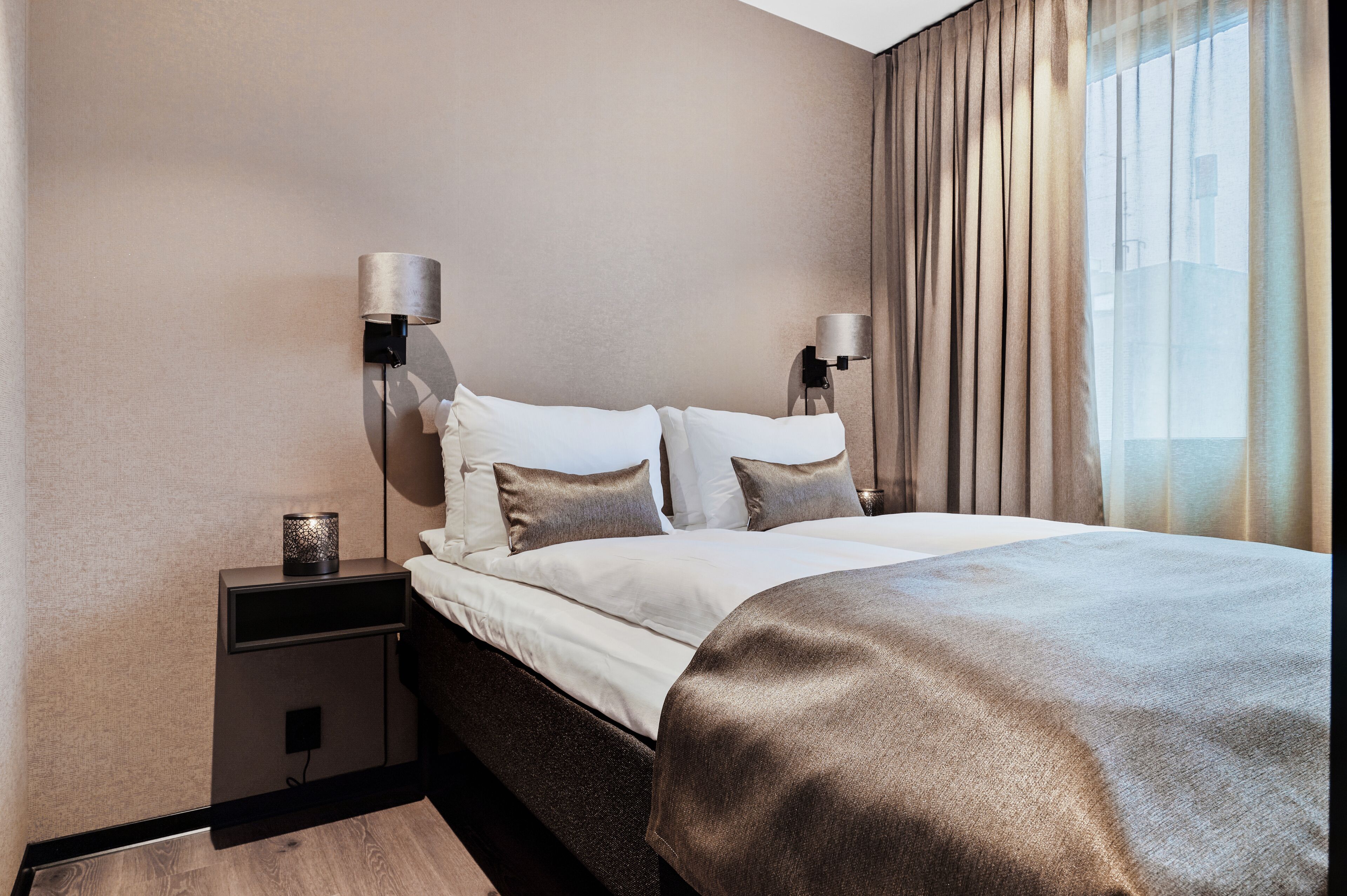 Une chambre d’hôtel moderne, avec un lit soigneusement fait, deux lampes de chevet et une vue sur la ville grâce à de grandes fenêtres.
