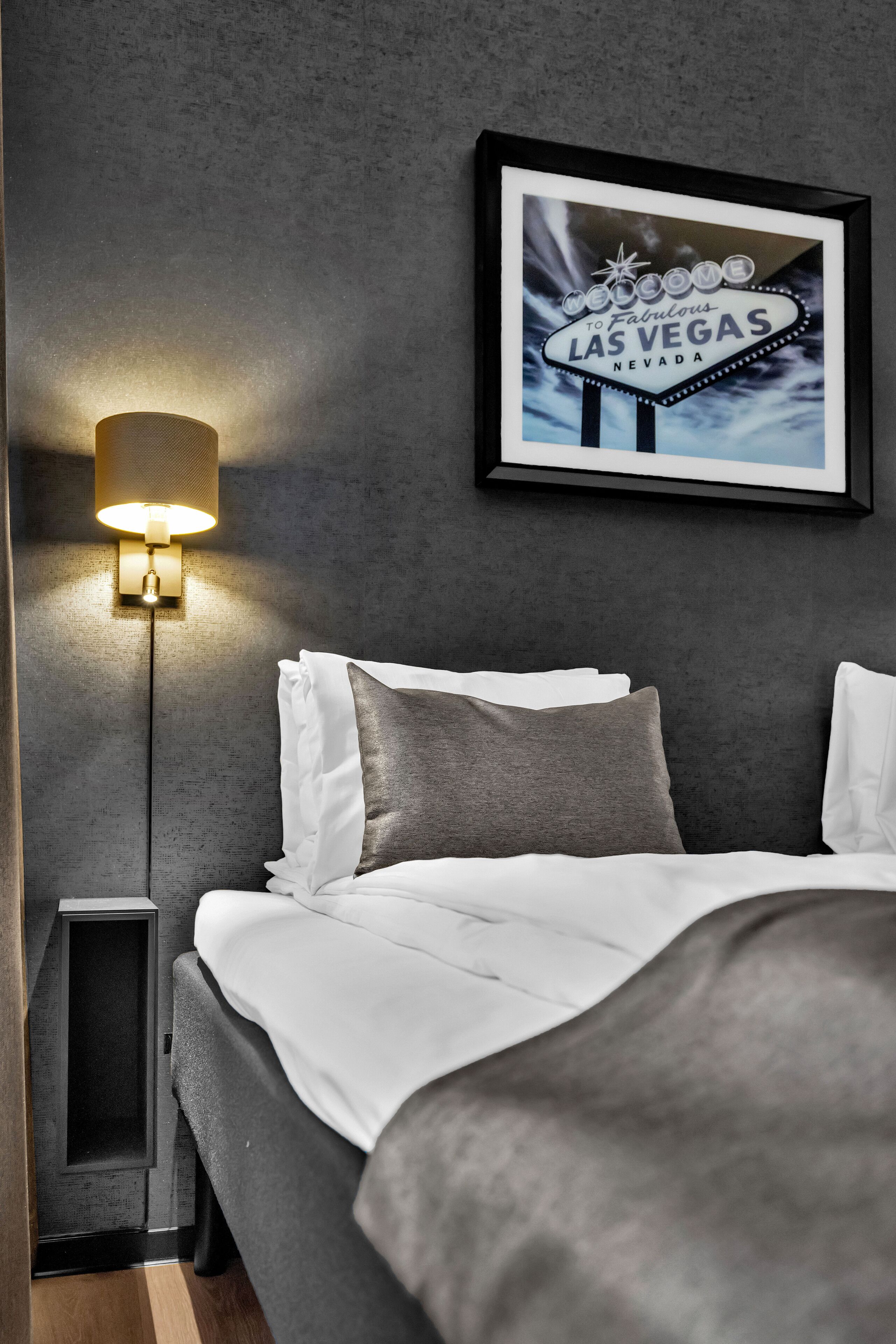 A neatly made bed with white and gray bedding, a wall-mounted framed sign reading "Welcome to Fabulous Las Vegas Nevada," and a floor lamp with a warm light.