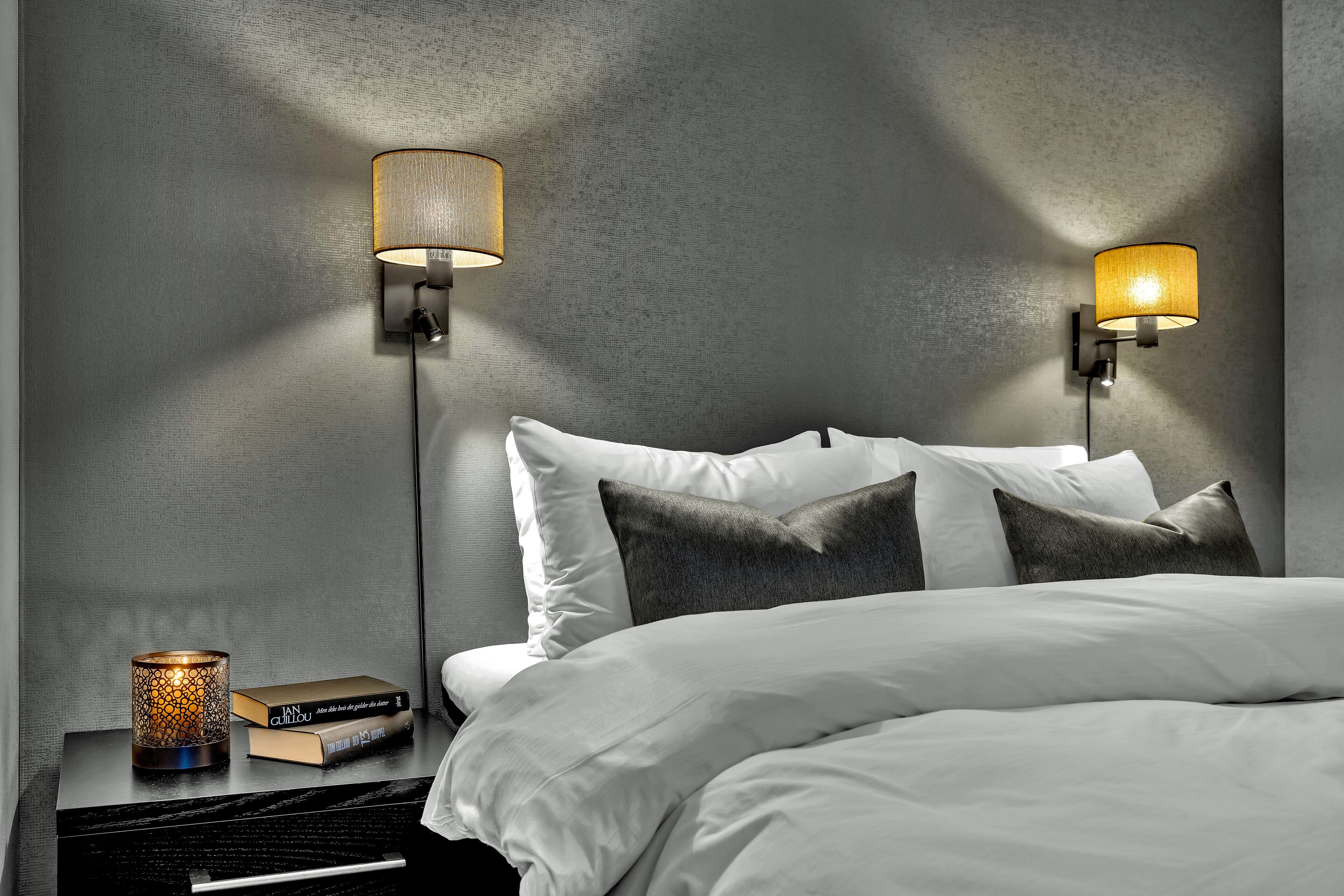 A neatly made bed with white linens and dark pillows, two wall-mounted lamps with cylindrical shades, and a small table with a candle and books.