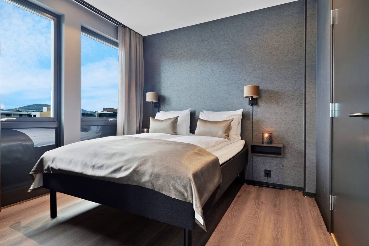 Una habitació d'hotel moderna amb un llit gran, làmpades de nit, una petita taula de nit i una vista a través de grans finestres.