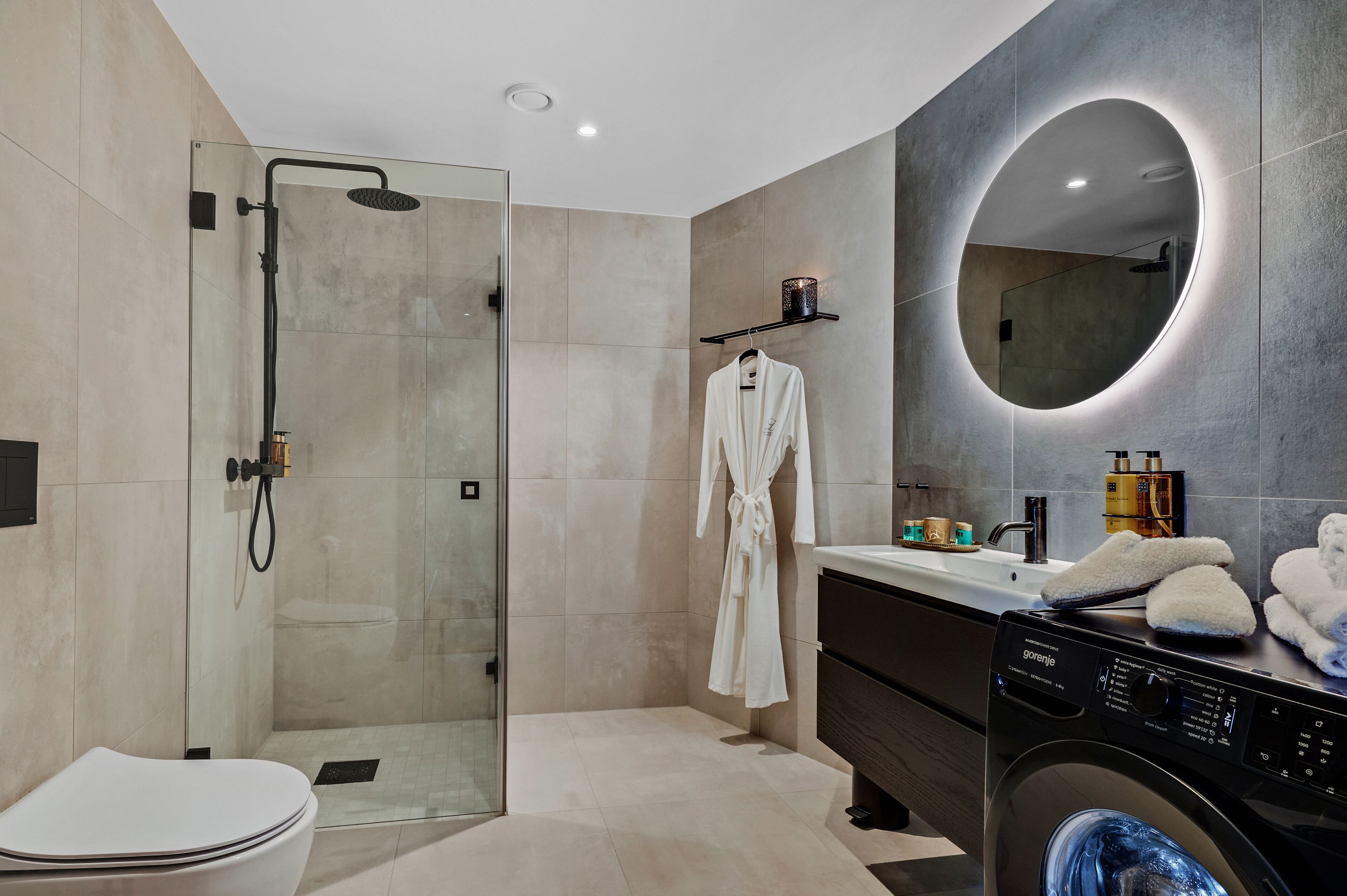 A modern bathroom with a glass-enclosed shower, a round mirror, a white robe, and a washing machine.
