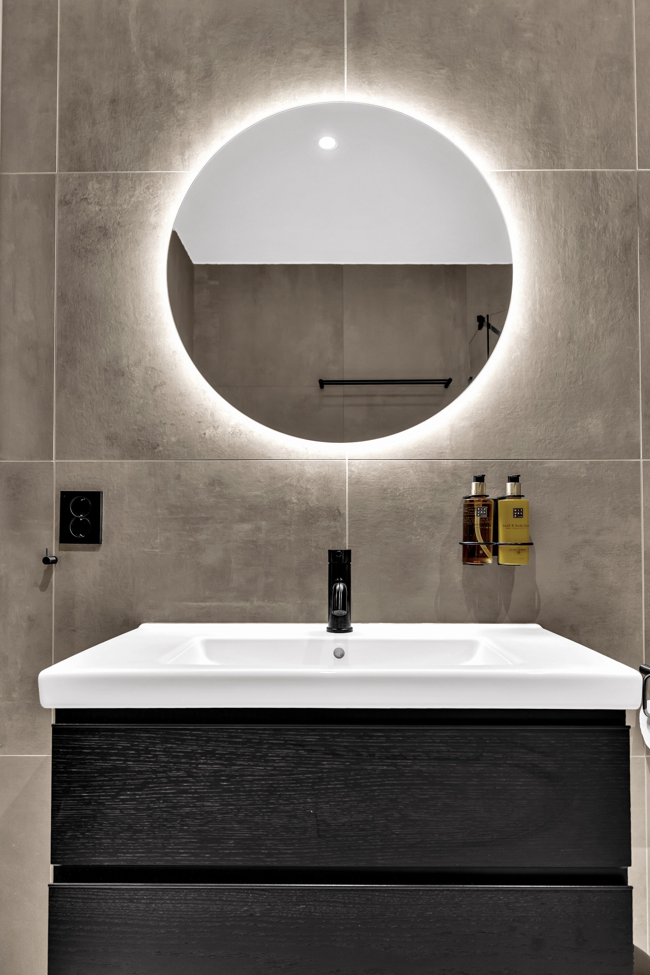 A modern bathroom with a round mirror, white sink, and dark wood cabinet.