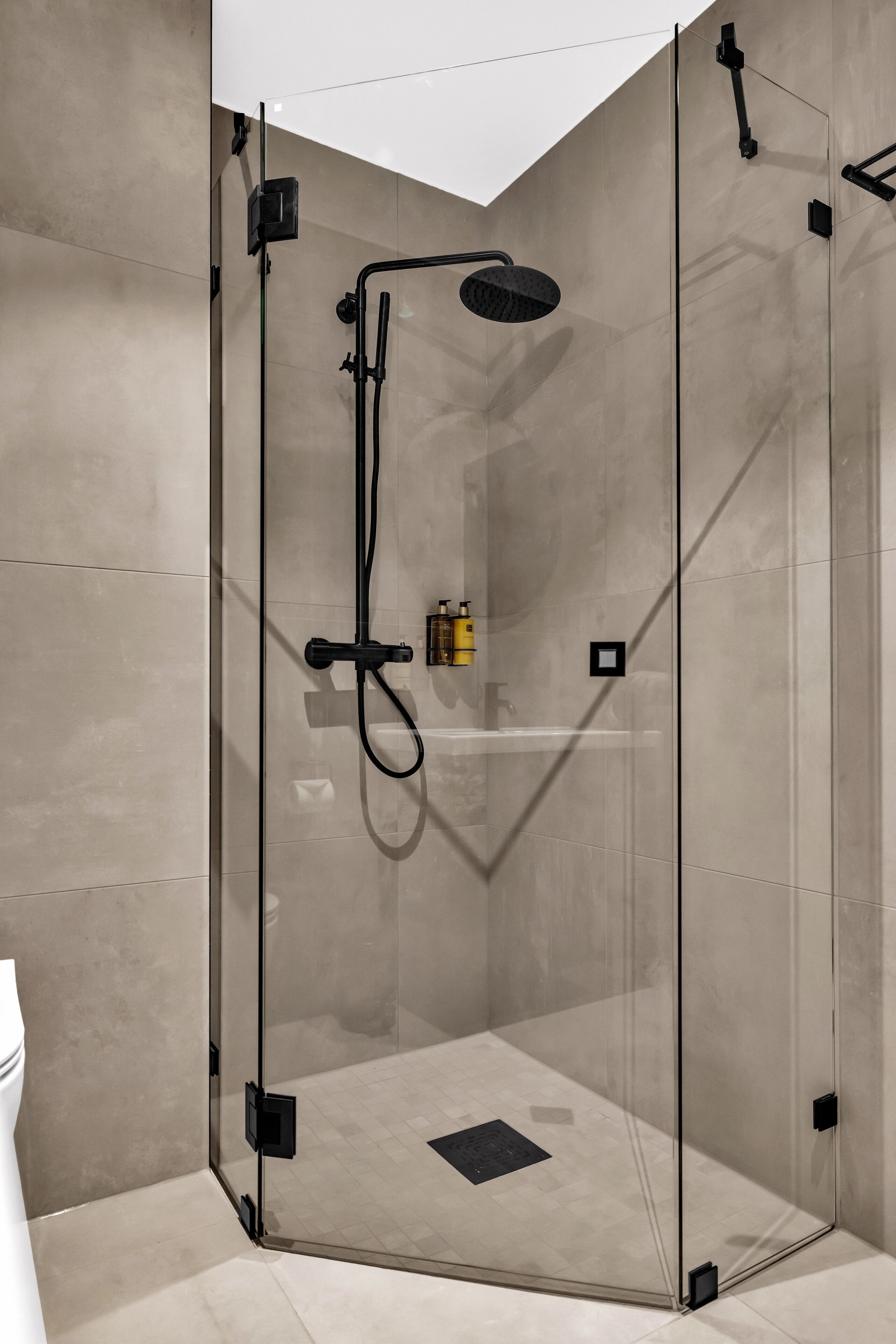 A glass-enclosed shower with a handheld showerhead, a wall-mounted control panel, and a small shelf with toiletries.