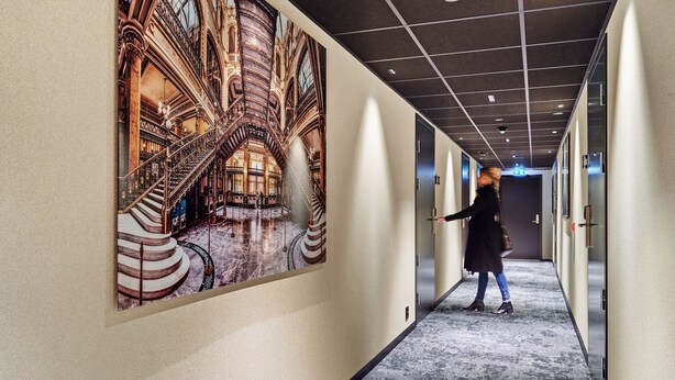 Un pasillo con un gran mural de un edificio histórico, suelo alfombrado y una persona caminando hacia una puerta.