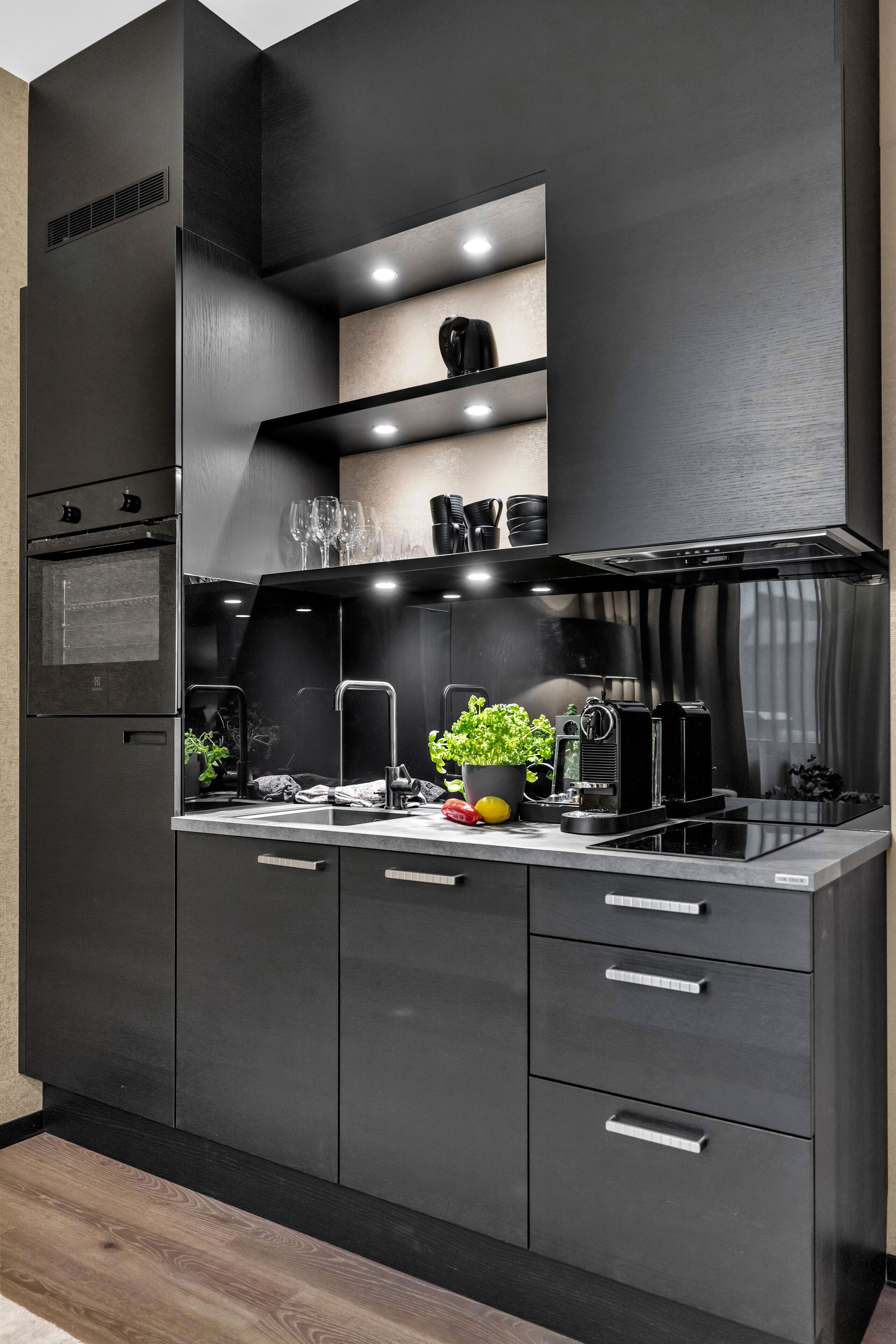 A modern kitchen with dark cabinetry, a built-in oven, a stainless steel range hood, and a countertop with a coffee machine and a potted plant.