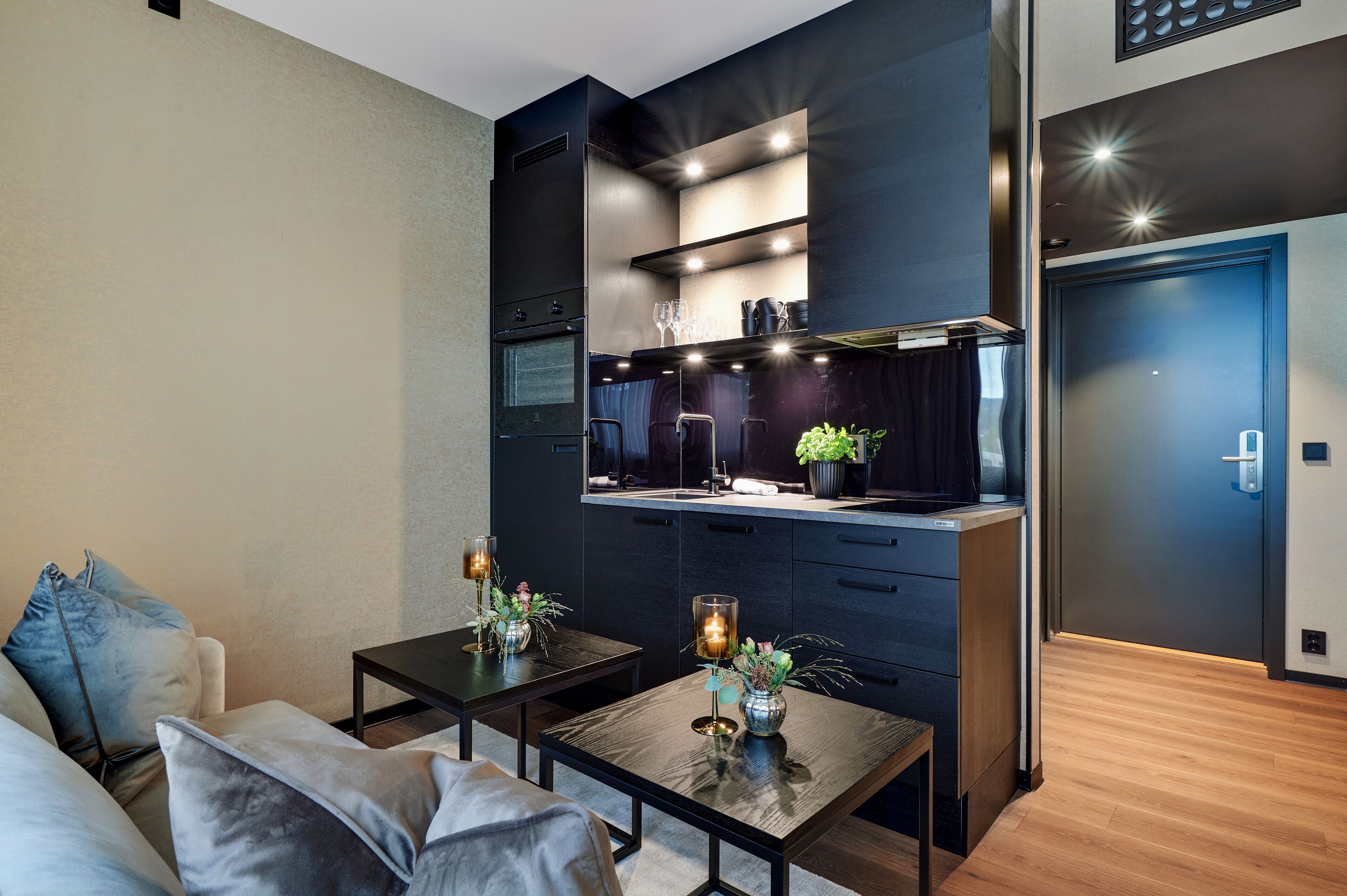 A modern kitchen with dark cabinetry, a built-in oven, and a countertop with a plant. There are two tables with candles and decorative items.