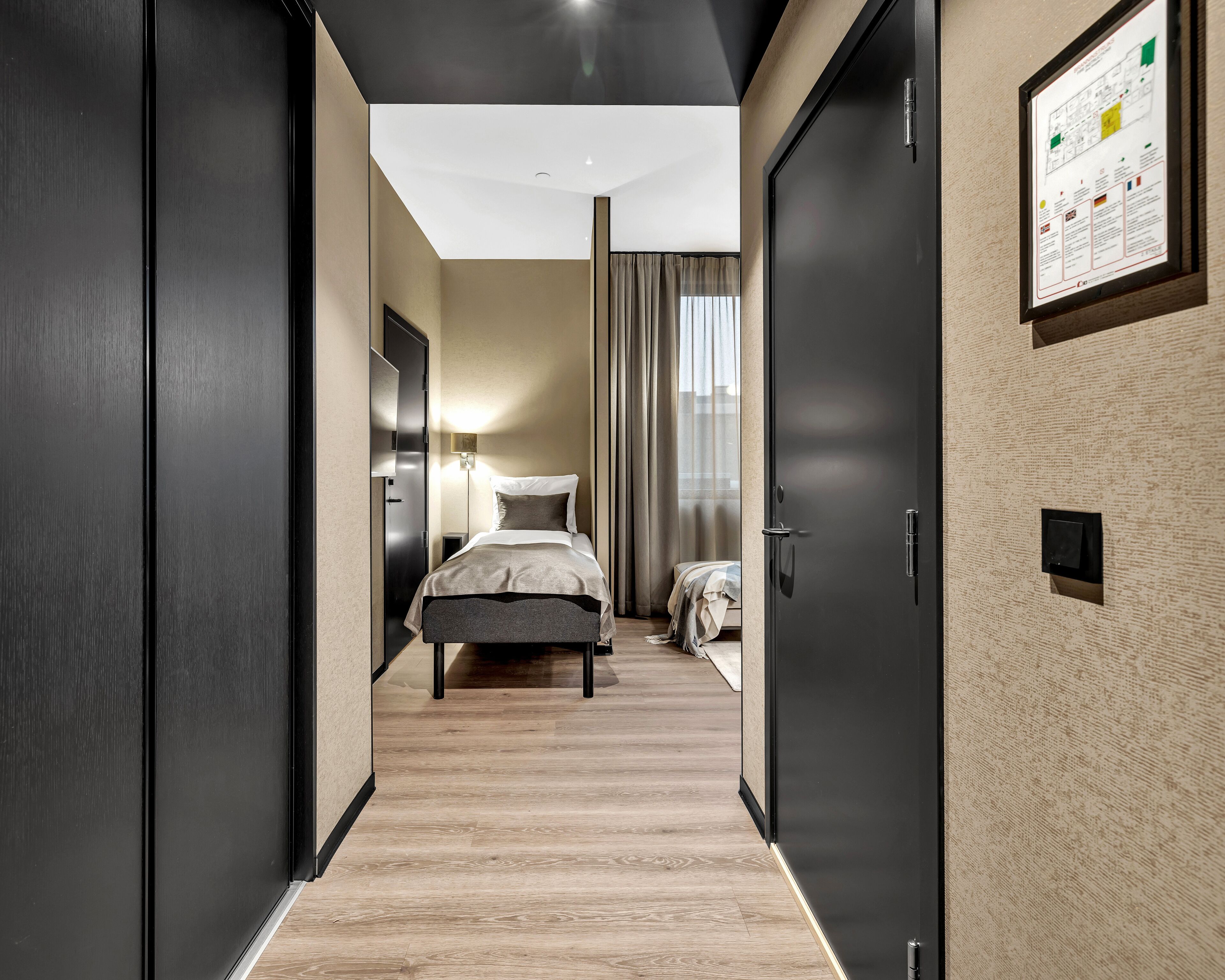 A modern hotel room with a wooden floor, a bed with a grey and white duvet, a nightstand with a lamp, and a framed map on the wall.