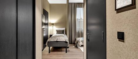 A modern hotel room with a wooden floor, a bed with a grey and white duvet, a nightstand with a lamp, and a framed map on the wall.