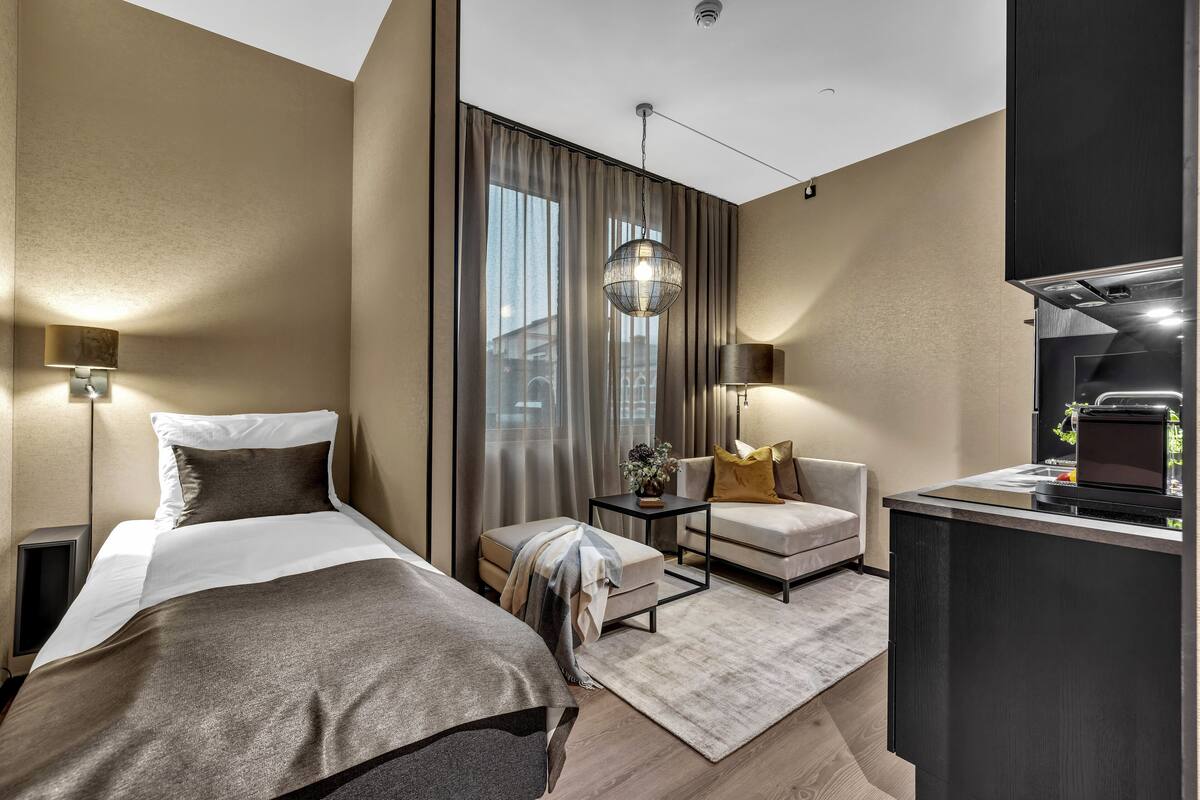 Una habitació d'hotel moderna amb un llit, una taula de treball, una cadira i una finestra gran amb cortines.