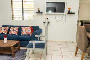 A living room with a blue sofa, a wooden coffee table, a TV mounted on the wall, and a small table with decorative items.