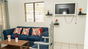 A living room with a blue sofa, a wooden coffee table, a white chair, a TV mounted on the wall, and a window with blinds. - Cozy house with 3 bedrooms, a 10-minute walk to the Samana boardwalk. (Samaná)