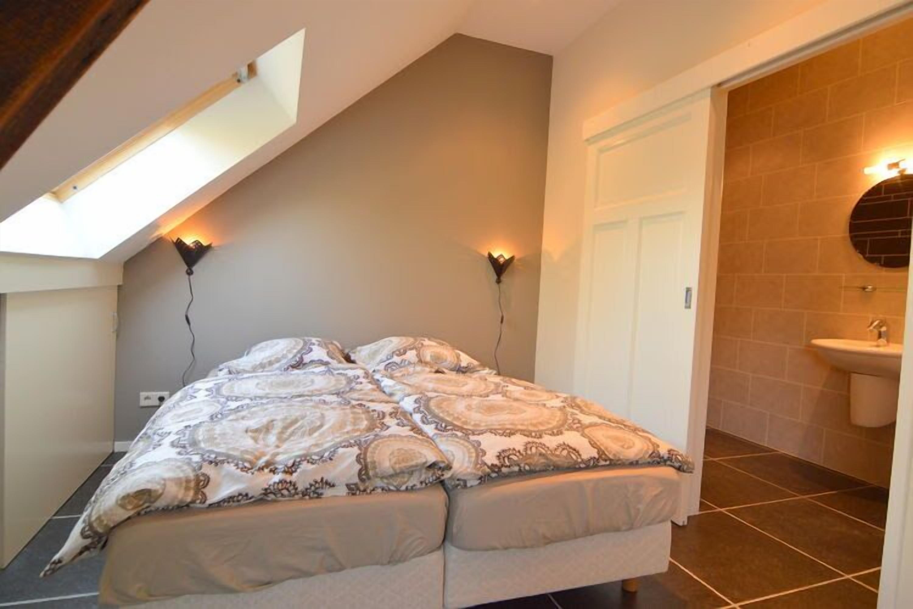 Un dormitorio con tragaluces, una cama con ropa de cama estampada, una lámpara de pared y un baño con lavamanos y paredes con azulejos.