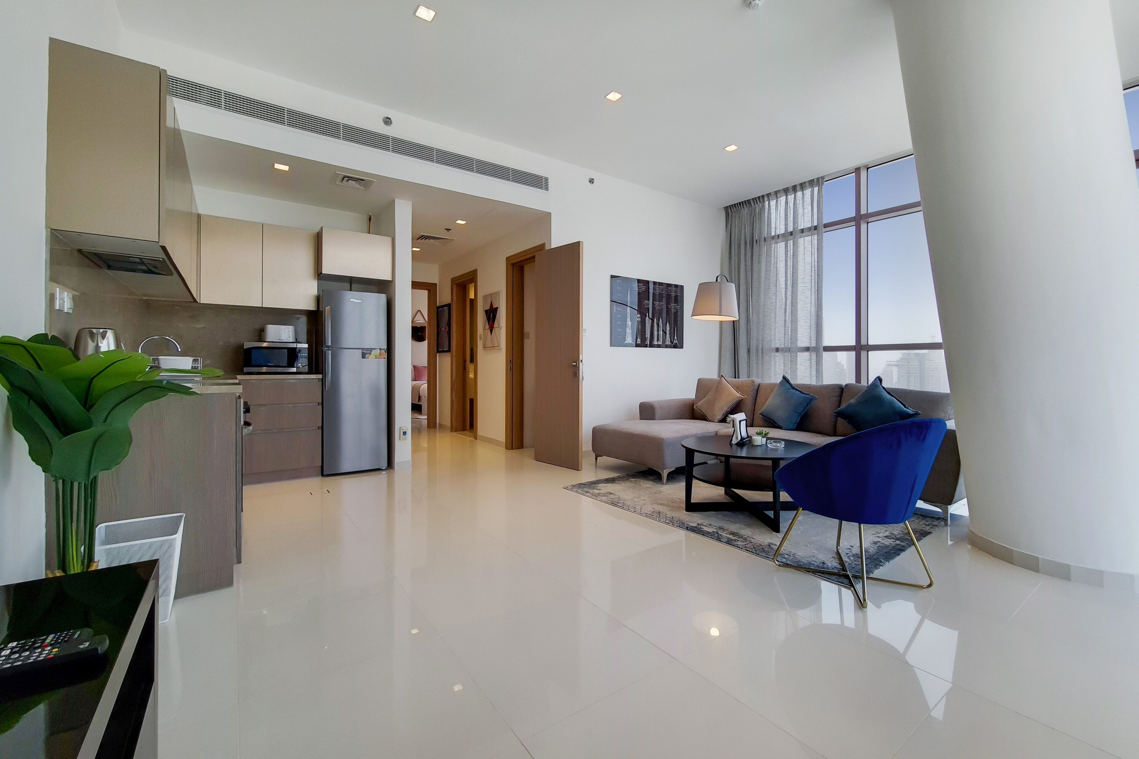 A modern living area with a sofa, coffee table, and a blue chair. There is a kitchen with a refrigerator and microwave.