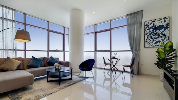 A modern living room with a sofa, coffee table, and chairs, large windows with curtains, and a painting on the wall.