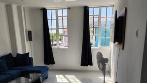 Un salon moderne avec un canapé bleu, une table basse en verre et un ventilateur sur pied. De grandes fenêtres laissent entrer la lumière naturelle et offrent une vue sur l’extérieur.
