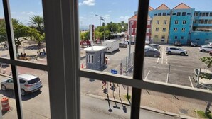 Une vue d’un parking avec plusieurs voitures, un petit bâtiment et, au fond, des bâtiments aux couleurs vives.