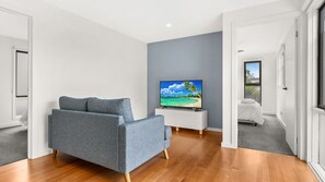 A modern living room with a grey sofa, a flat-screen TV on a white stand, wooden flooring, and a view of a bedroom with a bed and window. - The Bight - Cape Woolamai (Cape Woolamai)