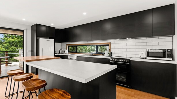 Una cucina moderna con mobili neri, un piano di lavoro bianco, una grande isola con sgabelli, un frigorifero, un fornello, un forno a microonde e una finestra con vista su piante.