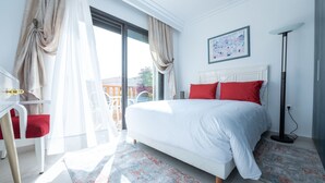 A modern bedroom with a large bed, white headboard, and red pillows. There is a desk with a chair, a floor lamp, and a window with sheer curtains. - Beautiful Villa  with heated pool Agadir! (Agadir)