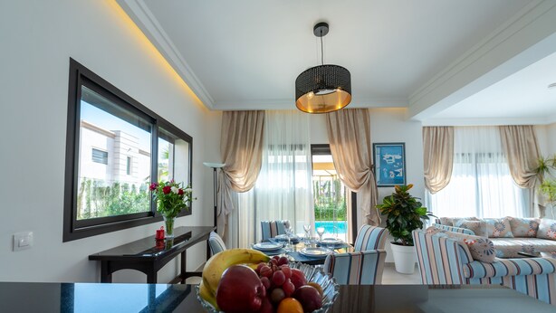 A modern living room with a dining area, featuring a large window, a dining table set with plates and glasses, a sofa with striped cushions, and a bowl of fruit on the coffee table. - Beautiful Villa  with heated pool Agadir! (Agadir)