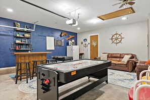 A room with a foosball table, a bar area, a brown leather sofa, and a ceiling fan.