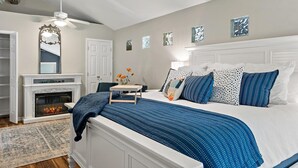 A bedroom with a bed, a fireplace, a ceiling fan, and artwork on the walls.
