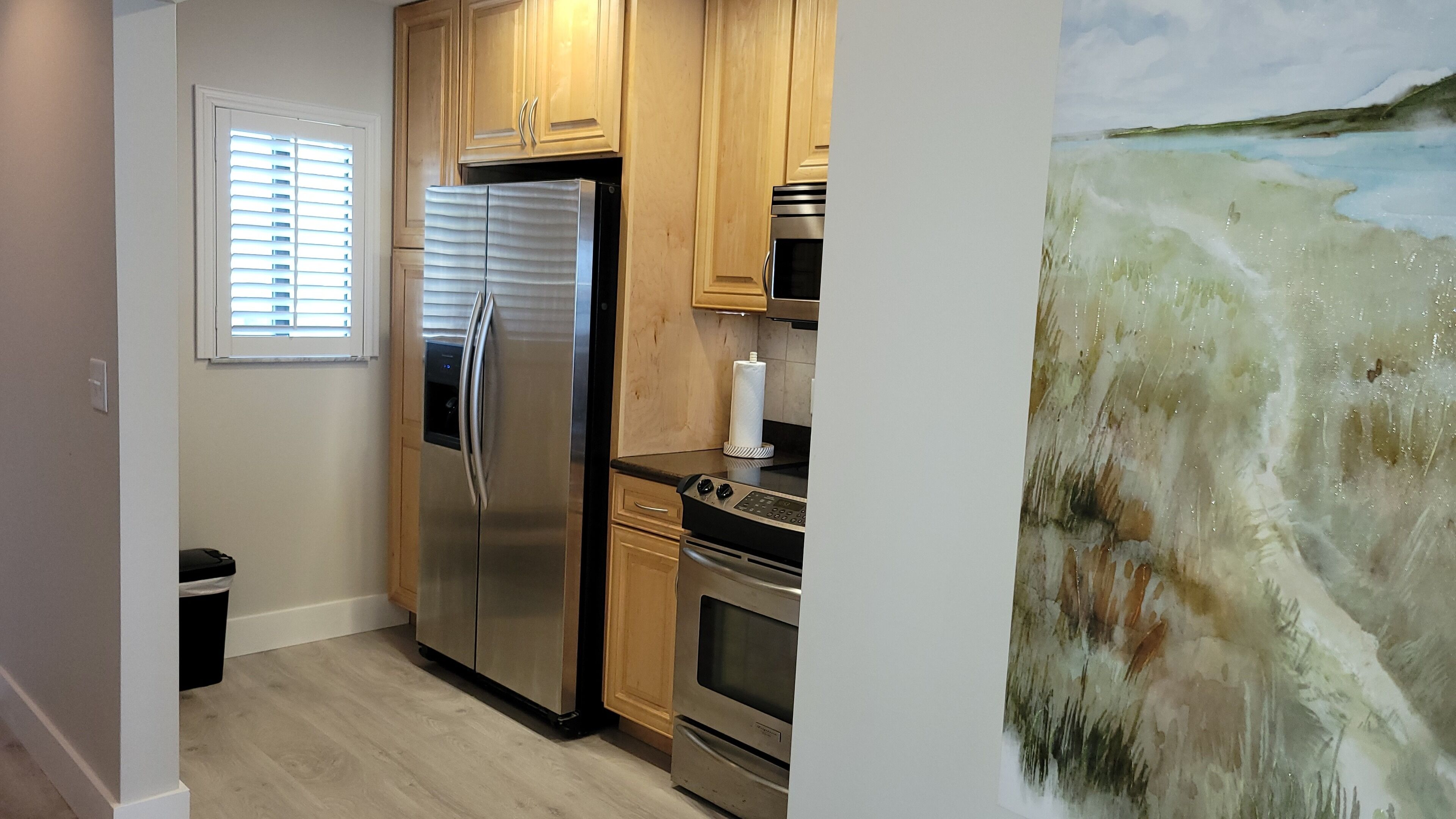 A kitchen with a stainless steel refrigerator, oven, and microwave. There is a window with blinds and a wooden cabinet.