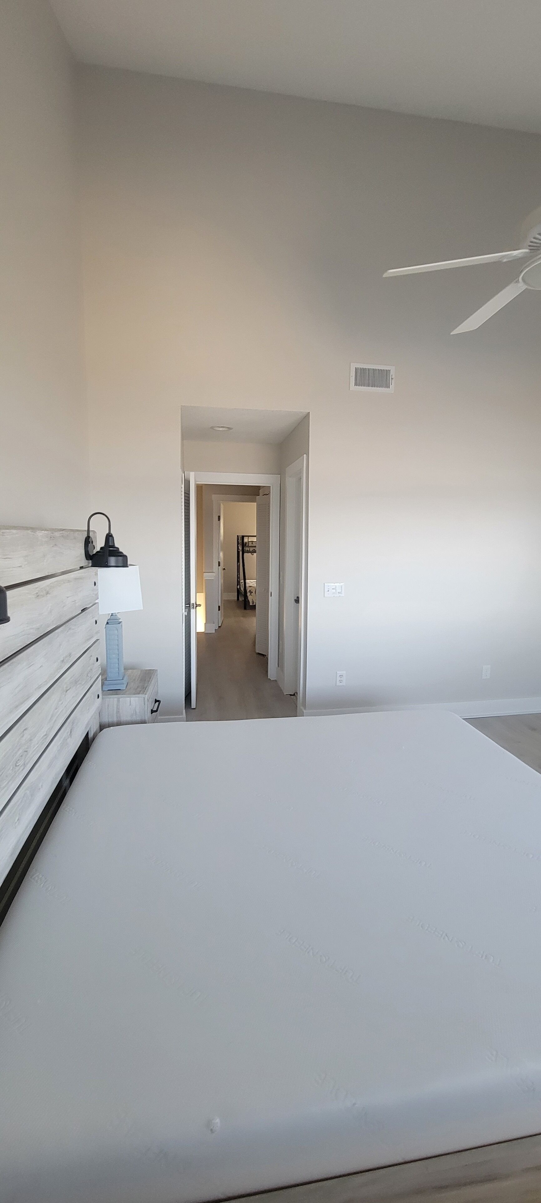 A bedroom with a bed, a nightstand, a lamp, and a ceiling fan. There is a hallway leading to another room.